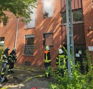 Das Bild zeigt Feuerwehr-Angehörige mit Atemschutzgeräten vor einem mehr geschossigen Ziegelhaus, aus dessen Fenstern weißer Rauch quillt