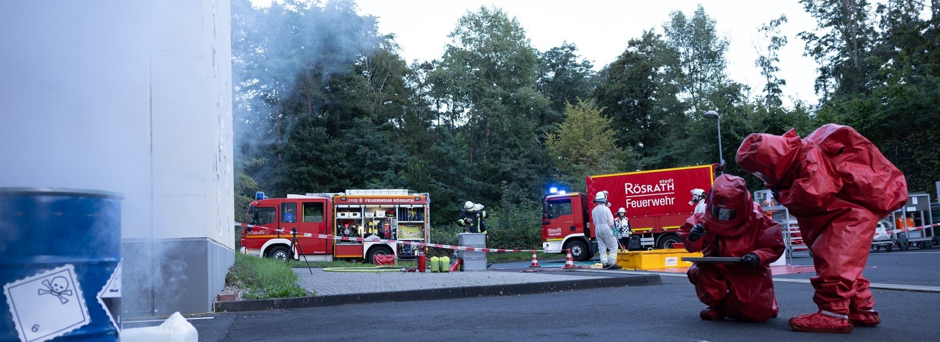 Im Vordergrund steht links ein blaues Fass mit einem Totenkopfsymbol, recht davon hocken zwei Personen in roten Chemikalienschutzanzügen. Im Hintergrund stehen zwei rote Feuerwehrfahrzeuge.