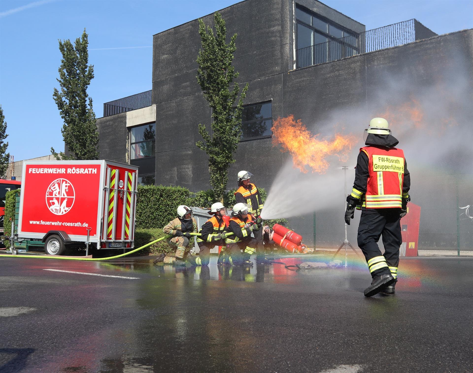 Vier Feuerwehrangehörige bekämpfen mit einem Schlauch und einem breiten Wasserstrahl eine Stichflamme, rechts im Bild steht ein einzelner Feuerwehrangehöriger mit einer roten Weste, links ein roter Anhänger mit der Aufschrift Feuerwehr Rösrath
