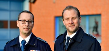 Das Bild zeigt zwei Männer in schwarzen Jacken vor einer orangenen Hauswand