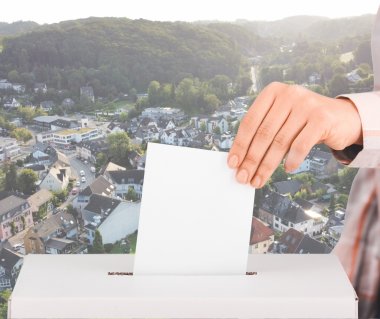 Das Bild zeigt den Einwurf eines Stimmzettels in eine Wahlurne vor Rösrather Kulisse.